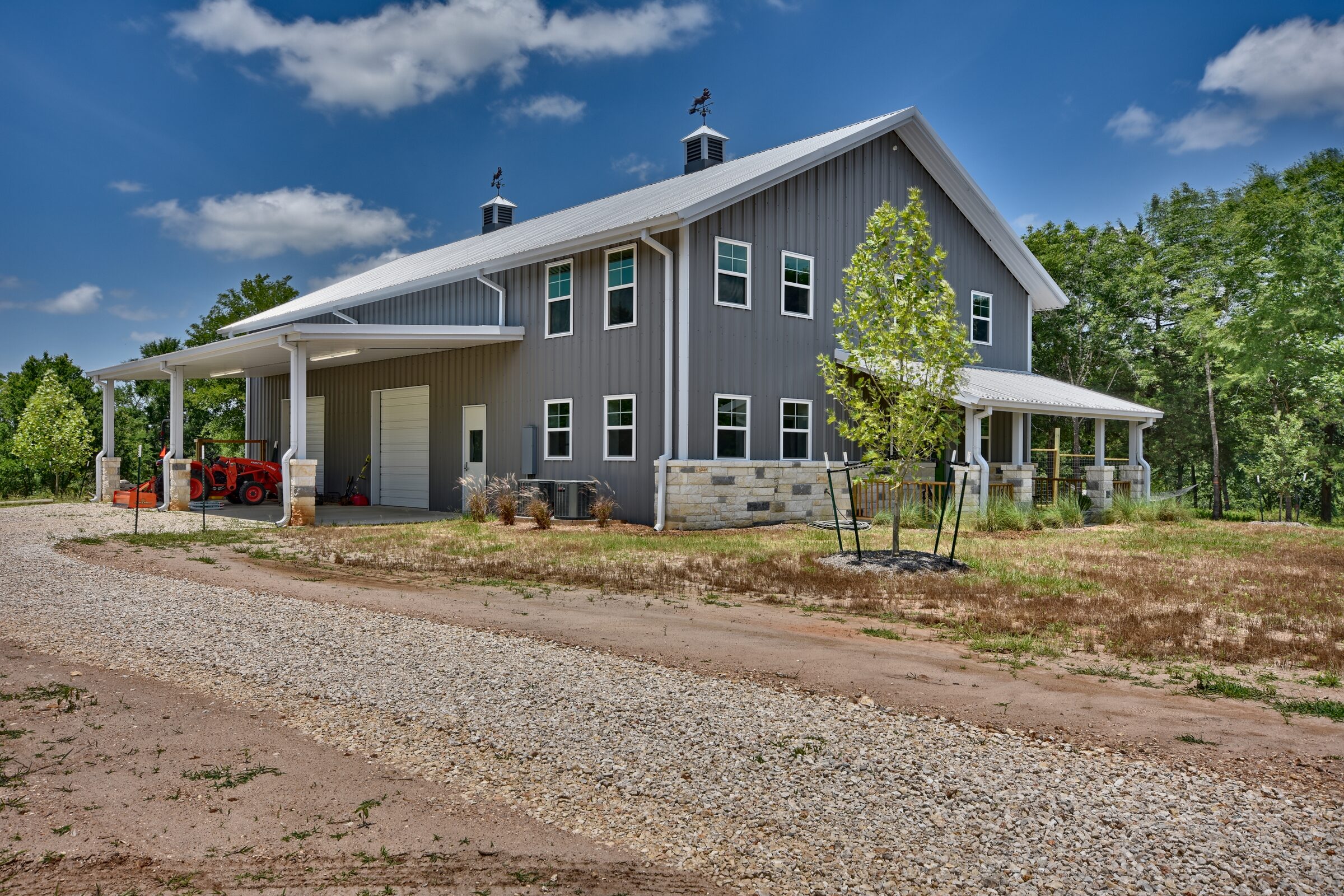 Grey two-storey barndominium with white trim, stone wainscot, cupolas and covered porches, with an open shop bay at one end
