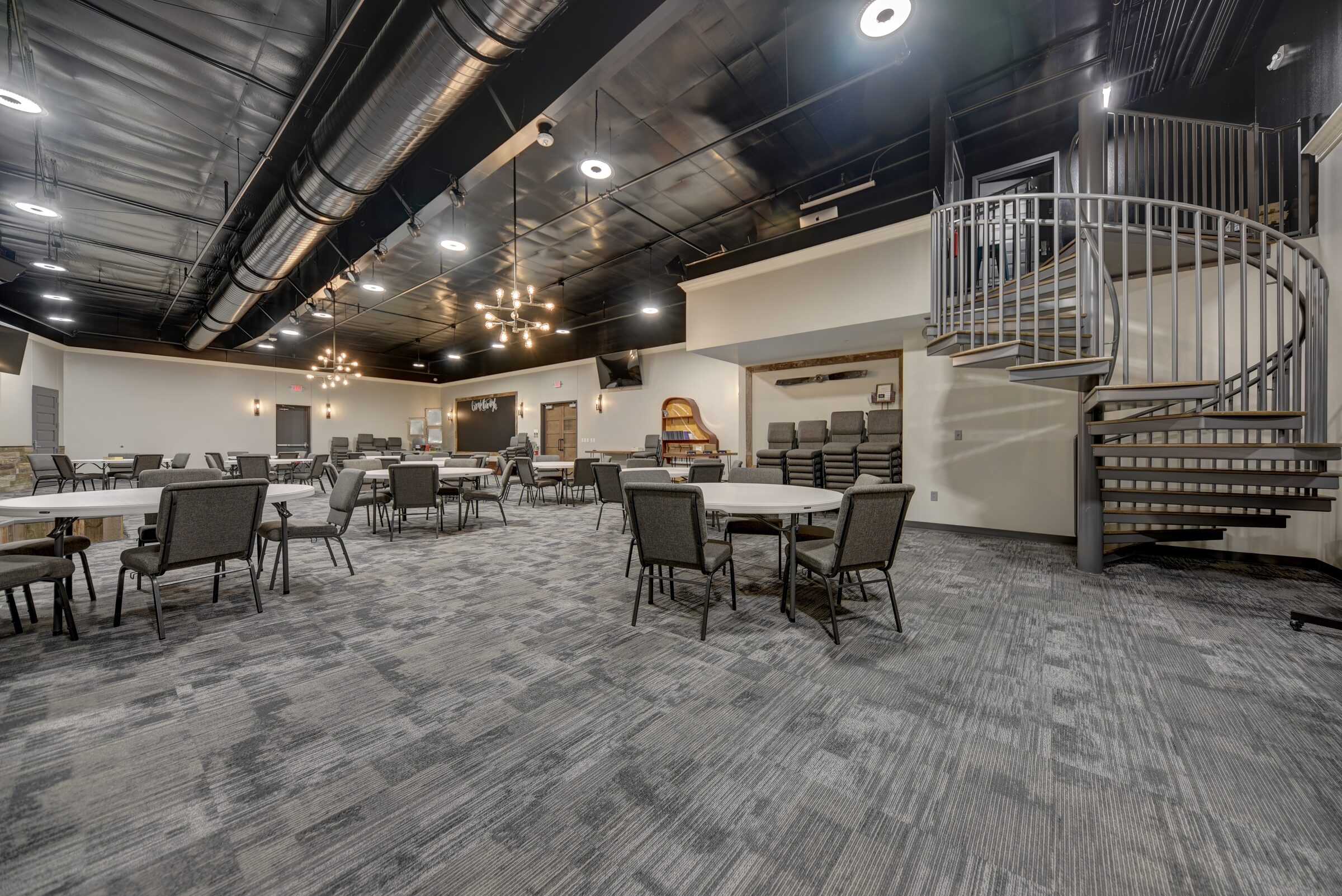 Finished interior hall with a spiral staircase, exposed black ceiling, chandeliers and round tables